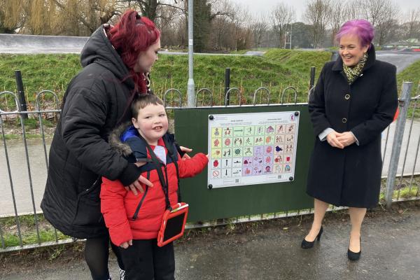 Reenie and Bash, a 6-year-old non-verbal boy, using a colourful symbol board in a local park to communicate and make a new friend, with the Minister for Children and Social Care observing nearby.